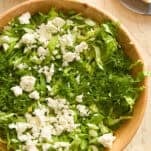 A wooden bowl filled with chopped lettuce, cucumber, fresh dill, and crumbled feta cheese. Nearby are a glass container of salad dressing, a plate with more feta, and a purple flower on a light surface.