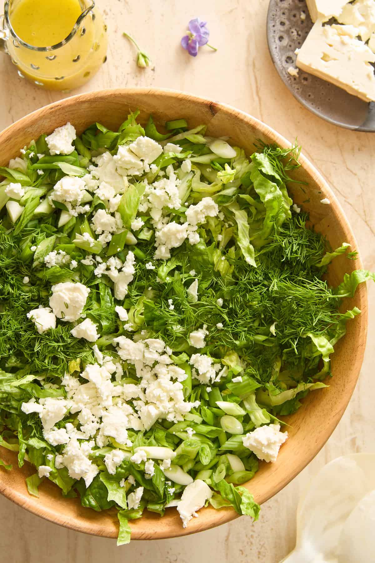 A wooden bowl filled with chopped lettuce, cucumber, fresh dill, and crumbled feta cheese. Nearby are a glass container of salad dressing, a plate with more feta, and a purple flower on a light surface.