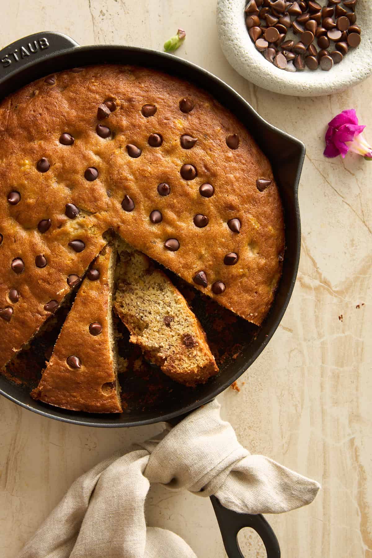 A skillet of chocolate chip banana bread with two slices cut, showing the soft interior. A beige cloth is wrapped around the pan handle, and a small bowl of chocolate chips and a flower are nearby on a light surface.