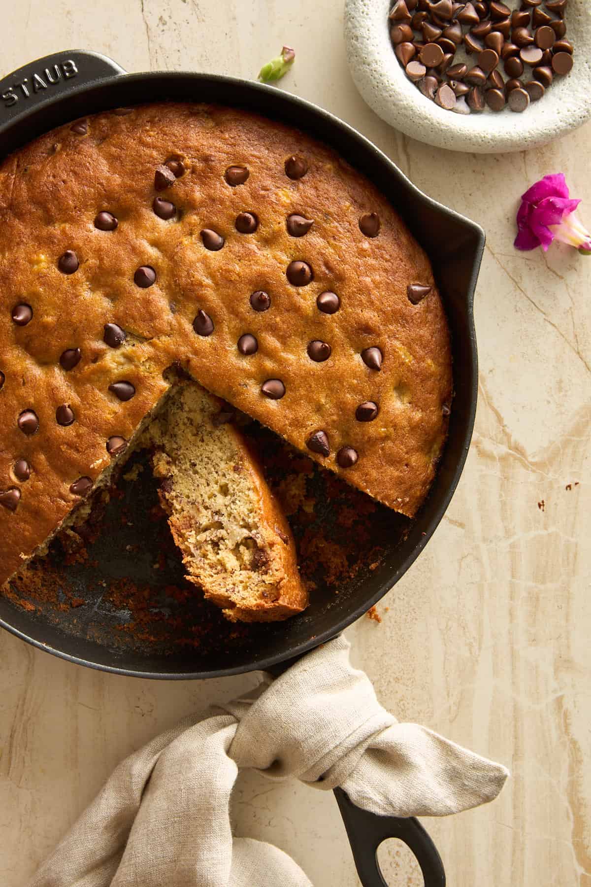 A skillet filled with chocolate chip banana bread, with one wedge sliced out. A beige cloth is tied around the skillet handle, and a small bowl of chocolate chips and a pink flower are nearby on a light countertop.