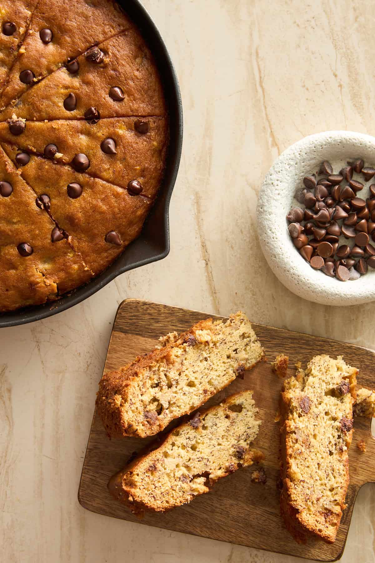A skillet of chocolate chip bread sits on a light counter. Two sliced pieces rest on a wooden board nearby, and a small bowl of chocolate chips is placed beside them.