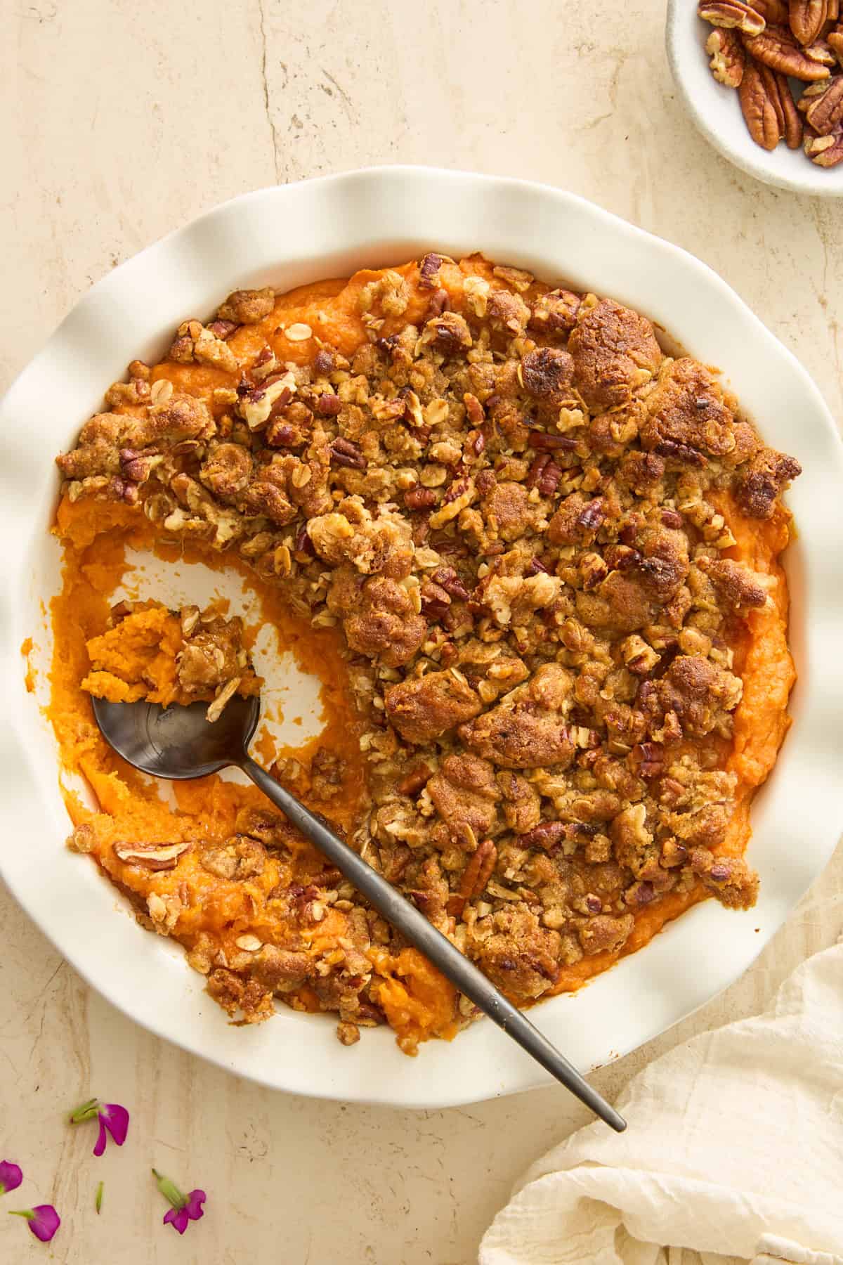 A white pie dish filled with sweet potato casserole topped with a golden-brown pecan crumble, with a silver serving spoon and some casserole scooped out. Pink flower petals and a bowl of pecans are nearby on a beige surface.