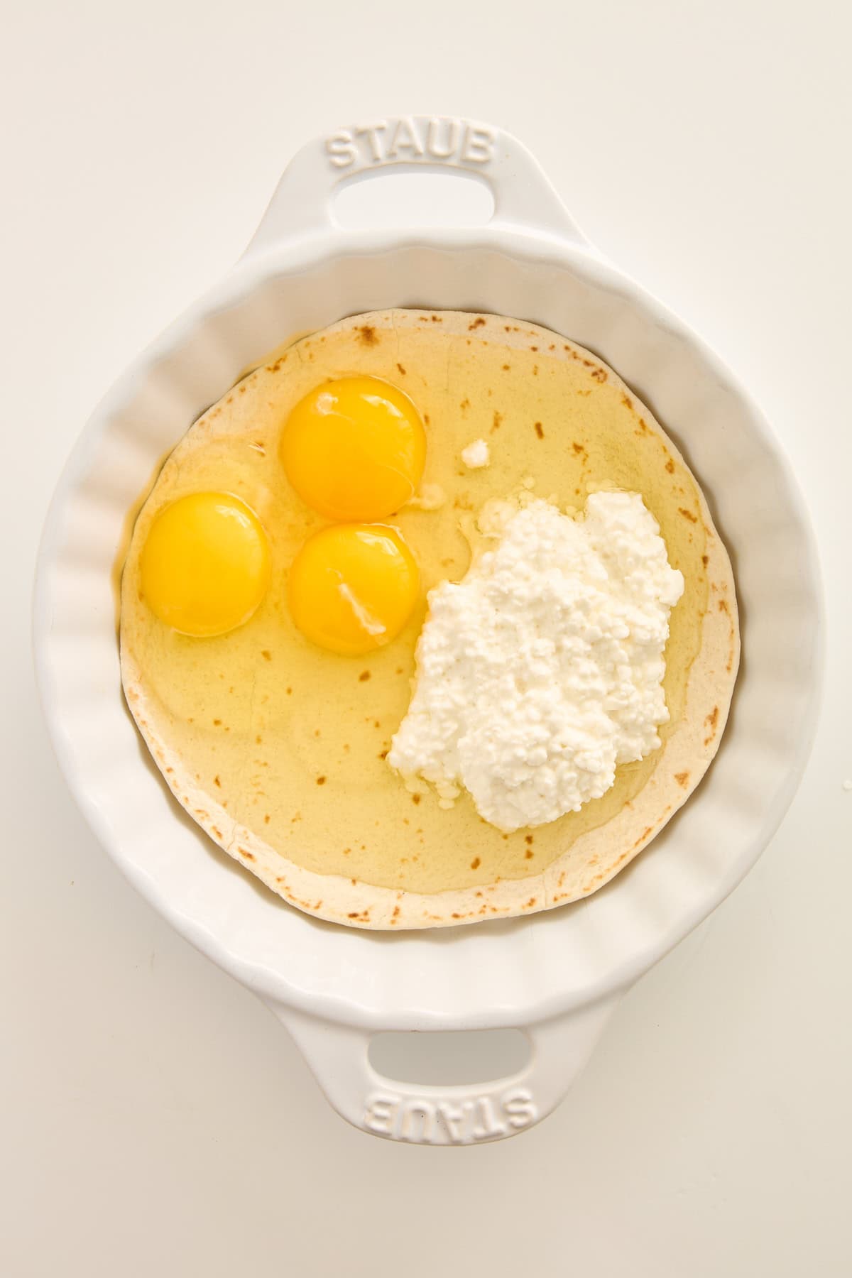 A white Staub baking dish containing three raw eggs, a heap of cottage cheese, and a creamy liquid mixture on top of a tortilla.