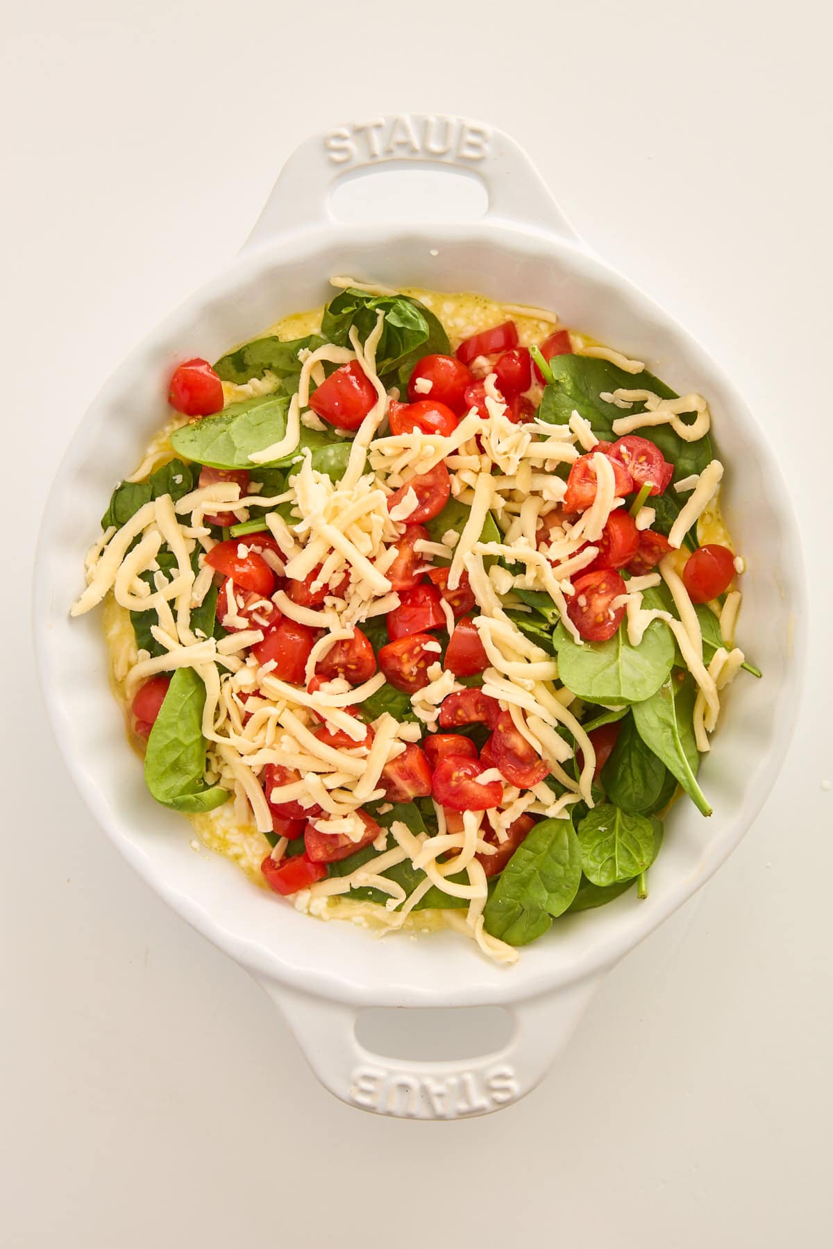 A white baking dish filled with fresh spinach, cherry tomato halves, shredded cheese, and beaten eggs, ready to be baked. The dish is on a plain white background.