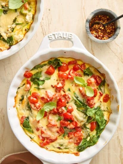 A baked frittata in a round white dish, topped with cherry tomatoes, spinach, melted cheese, and fresh basil leaves. A small bowl of red pepper flakes with a spoon is nearby on a light countertop.