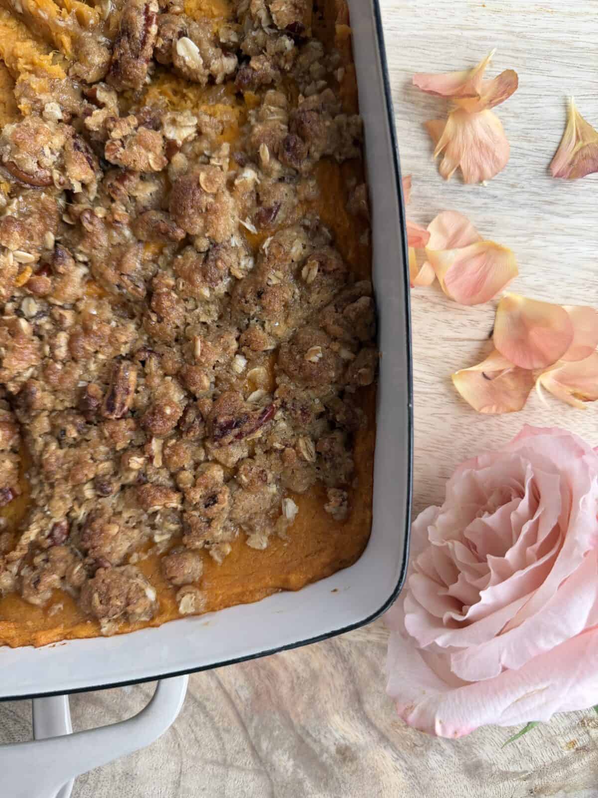 A close-up of a casserole dish filled with sweet potato casserole topped with a crumbly pecan oat streusel. Next to the dish are pink rose petals and a single large pink rose on a wooden surface.