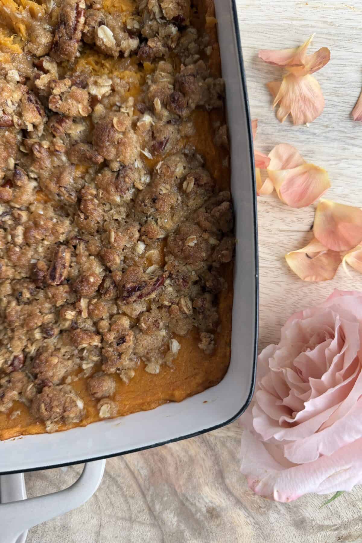 A close-up of a casserole dish filled with sweet potato casserole topped with a crumbly pecan oat streusel. Next to the dish are pink rose petals and a single large pink rose on a wooden surface.