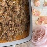 A close-up of a casserole dish filled with sweet potato casserole topped with a crumbly pecan oat streusel. Next to the dish are pink rose petals and a single large pink rose on a wooden surface.