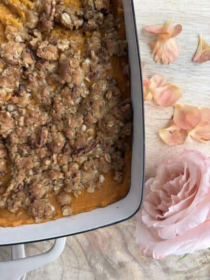 A close-up of a casserole dish filled with sweet potato casserole topped with a crumbly pecan oat streusel. Next to the dish are pink rose petals and a single large pink rose on a wooden surface.