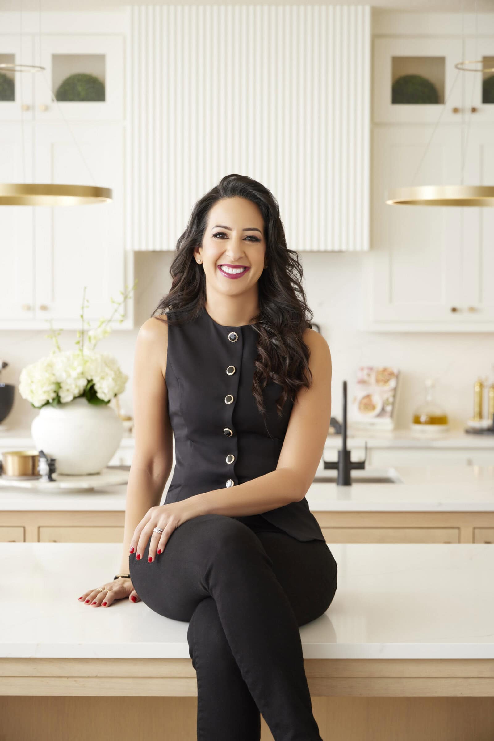 Alia Elkaffas with long dark hair and a sleeveless black top sits on a kitchen counter, smiling. The modern kitchen background includes white cabinets, hanging lights, and a vase of white flowers.