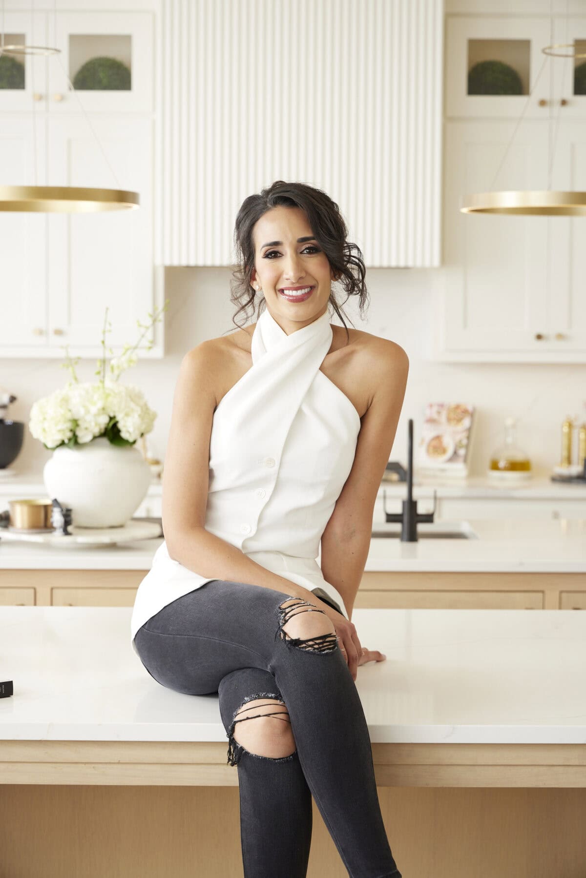 Radwa Elkaffas with dark hair in an updo, wearing a white halter top and ripped black jeans, sits on a modern kitchen counter, smiling. Behind her are white cabinets, flowers, and kitchen decor.