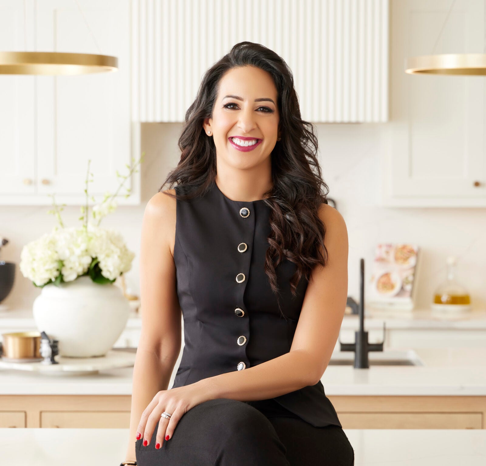 Alia Elkaffas with long dark hair and a sleeveless black top sits on a kitchen counter, smiling. The modern kitchen background includes white cabinets, hanging lights, and a vase of white flowers.
